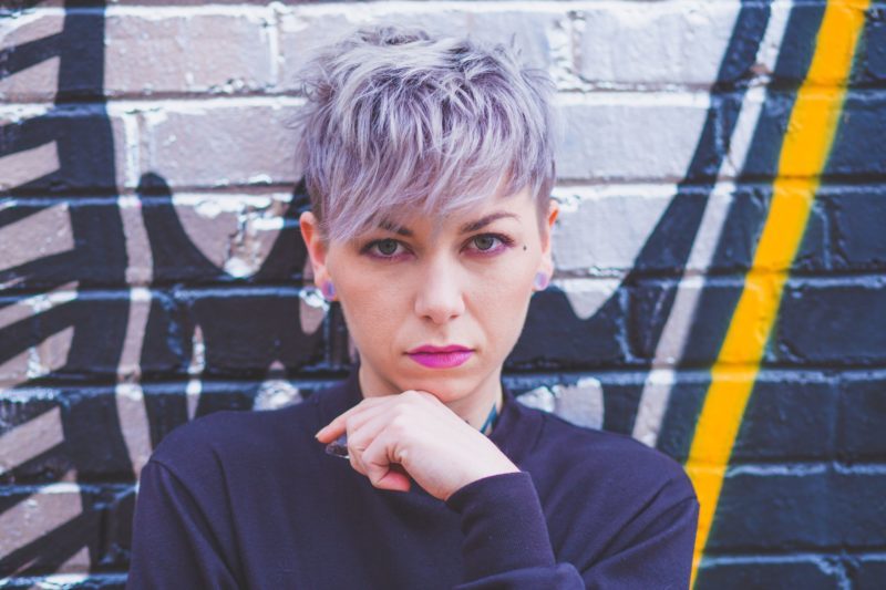 Person with pastel purple pixie hair standing in front of a graffited wall