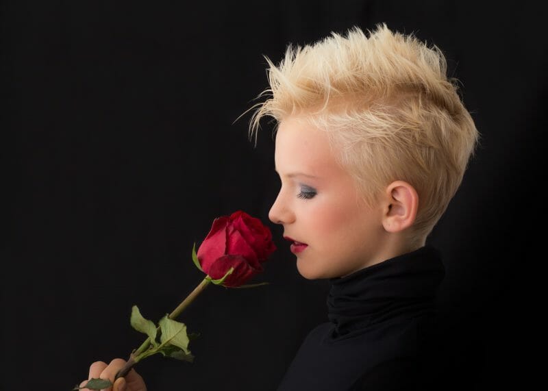 Person with a pixie cut smelling a rose on a black background